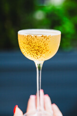 A hand holds a glass of golden bubbly liquid, possibly champagne, with tiny bubbles clinging to the side. The blurred green background adds a touch of luxury