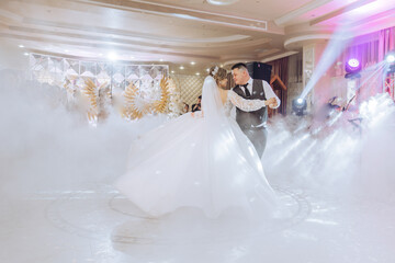 A bride and groom are dancing in a ballroom with a fog machine. The bride is wearing a white dress...
