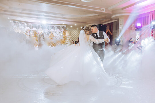 A bride and groom are dancing in a ballroom with a fog machine. The bride is wearing a white dress and the groom is wearing a suit. The fog machine creates a misty atmosphere, adding to the romantic