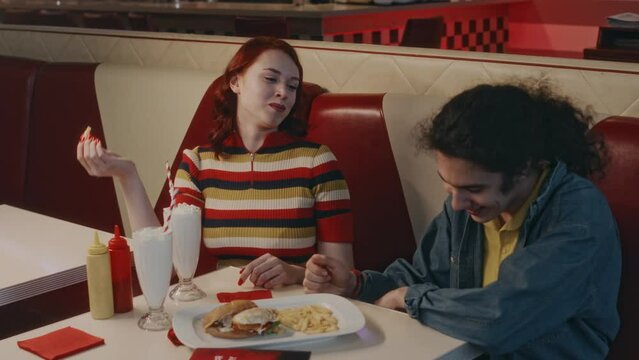 Medium shot of cheerful Caucasian lady feeding her boyfriend with fries and teasing him, eating it herself while on evening date in retro style cafe