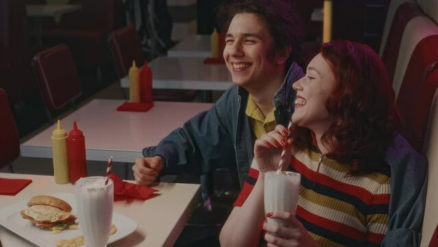 Side shot of talkative Caucasian guy telling amusing stories with hand on his girlfriends shoulder while she drinking milkshake, evening date in authentic retro diner