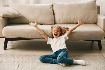 Joyful little girl sitting on the floor with arms outstretched in front of a cozy couch in a bright...