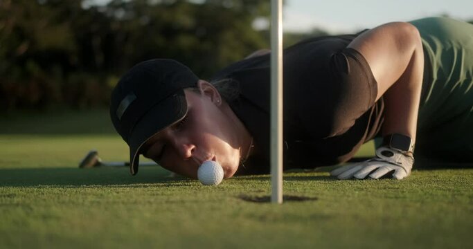 A young woman lies on a green lawn on the country club while playing golf and blows on a golf ball to hit the hole. She laughs and has fun. Trick during the game. Strategy on the path to victory.