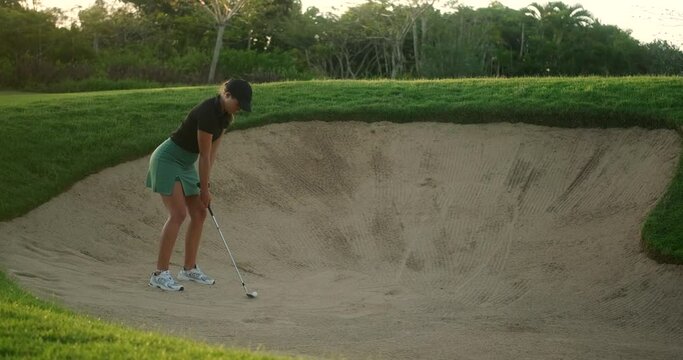 A young female professional golfer fell into a sand trap. She hits the ball from the bunker up the hill and jumps up from the bottom to see if she hits the hole. Making golf more difficult.