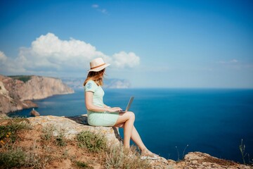 A woman is sitting on a rock overlooking the ocean with a laptop in front of her
