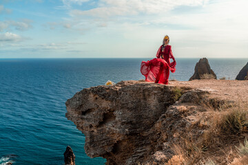A woman in a red dress stands on a rocky cliff overlooking the ocean. The scene is serene and...
