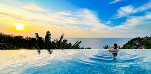 A woman enjoys a serene moment in an infinity pool overlooking the ocean at sunset. The lush...