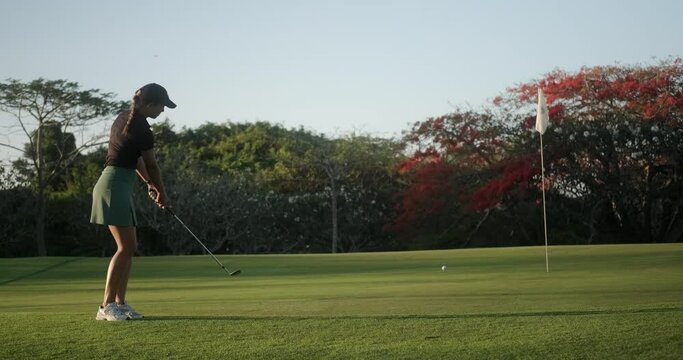 Young woman beginner golfer. She undergoes training and does not hit the ball in the hole. Upset woman is unhappy with her failure. She walks towards the ball and swings the golf club.
