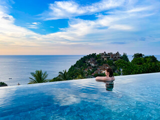 A woman enjoys a serene moment in an infinity pool overlooking the ocean at sunset. The lush tropical scenery and peaceful ambiance create a perfect setting for themes of relaxation, luxury, and © DmitriiArtamonov