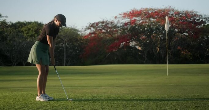 A young attractive woman professional golfer in a country club on a green lawn hits a ball into a hole with a golf club. She jumps and rejoiced at her victory in the competition. Happy winner.
