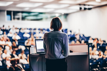 Female speaker giving a talk on corporate business conference. Unrecognizable people in audience at...