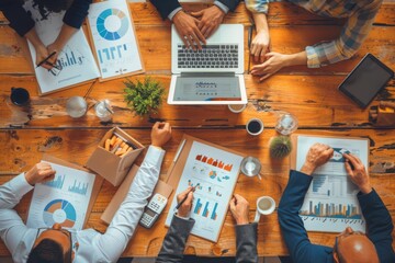 Image showing overhead view of a diverse team working together at a busy wooden table filled with laptops, documents, and hands pointing at charts