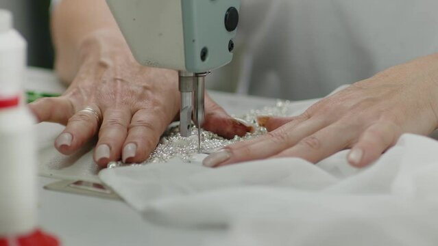  Fashion designer using a sewing machine at her workplace. Female seamstress sews white fabric items on a sewing machine. 