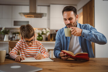 Father drink coffee and hold book while son do homework at home