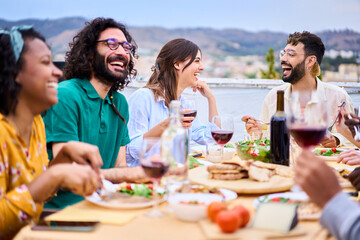 Millennial happy people laughing sitting at food table drinking red wine enjoying sunny free time together on rooftop. Group of young joyful friends celebrating barbecue meeting summer party outdoor
