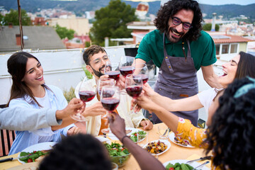 Joyful millennial group friends toasting glasses red wine celebrating rooftop barbeque summer party. Young people together enjoying cheers alcoholic drinks sitting at terrace lunch table outdoors 