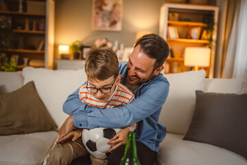 Adult father hug son who hold football ball while watch football match