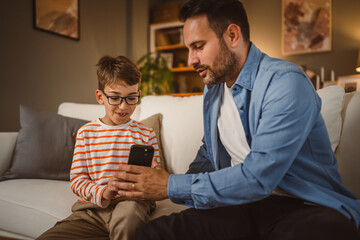 Father and son sit on sofa and use mobile phone at home have fun