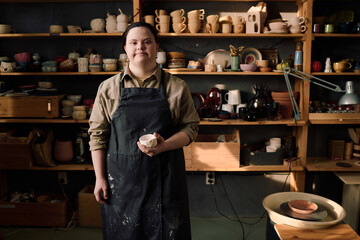 Medium long shot of young Caucasian female artisan with Down syndrome standing in pottery workshop looking at camera, copy space