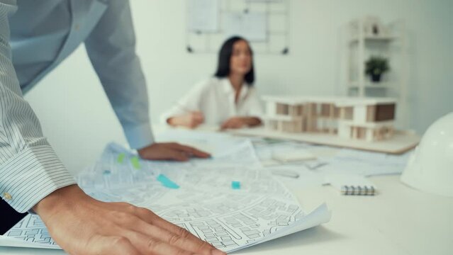 Skilled caucasian interior designer working with house model while discussing with asian architect engineer about house plan on meeting table with blueprint and house model scatter around. Immaculate.