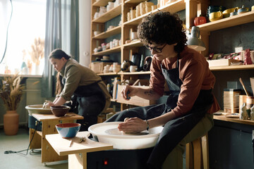 Two diverse women wearing aprons shaping clay on potters wheels during class in workshop, copy space