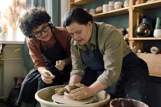 Cheerful Caucasian female artisan sitting next to woman with Down syndrome teaching her how to shape clay on potters wheel