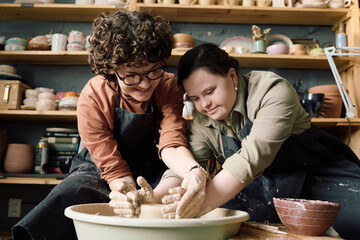 Low angle view shot of two diverse women wearing aprons having fun during pottery class shaping clay on wheel together