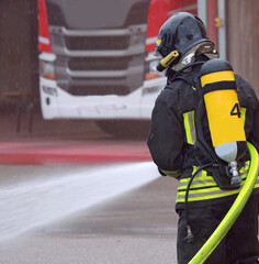 Firefighter with oxygen tank wearing protective helmet sprays a lot of foam jet while it rains