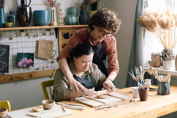 Modern artisan teaching young woman with Down syndrome how to roll out clay during pottery class in...