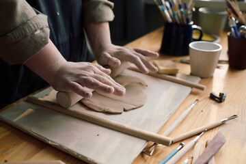 Closeup of hands of unrecognizable artisan rolling out piece of clay with use of rolling pin, copy...