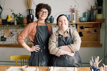 Portrait of two diverse female artisans wearing aprons standing at table in pottery workshop posing...