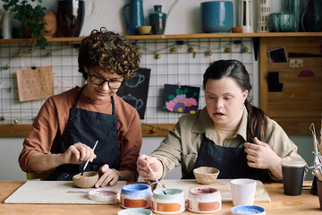 Two diverse Caucasian women attending pottery workshop sitting at table painting handmade ceramic bowls
