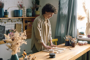 Modern Caucasian woman wearing casual clothes standing at table in pottery workshop rolling out piece of clay