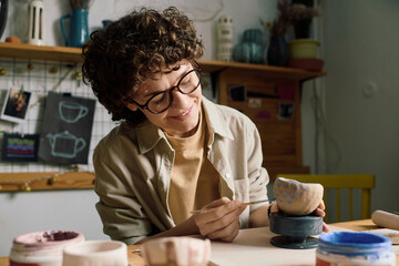 Fototapeta premium Cheerful Caucasian woman sitting at table in workshop painting blue hearts on handmade ceramic bowl