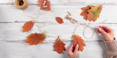 Homemade garland of autumn leaves. Womans hand hanging leaves on rope with clothespin on white wooden background. Home diy decor for celebrating fall holidays, Thanksgiving and Halloween banner