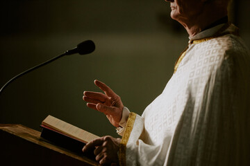 Side view crop shot of senior Caucasian Catholic priest standing at lectern with Bible book on it...