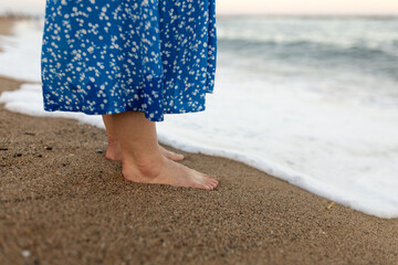 Woman in dress walking on sand beach. Wave motion coming to the foot, foaming sea texture. Summer and vacation concept.