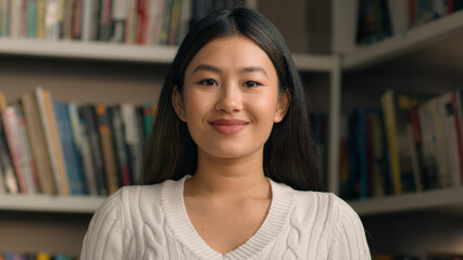 Young asian woman student posing in university library or bookstore with bookshelf in background...