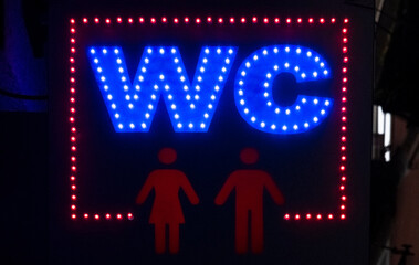 A colorful neon sign suspended on a wall in a dark alley at night, WC (stands for Water Closet, aka...