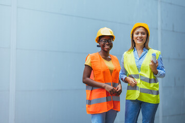 Two cheerful women in high-visibility vests and safety helmets stand against a blue wall, embodying...