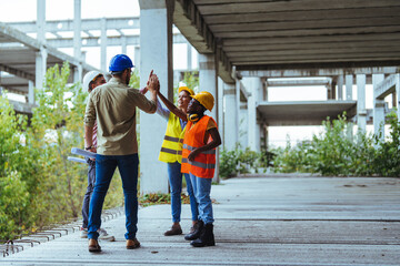 A joyful group of diverse construction workers exchange high-fives on a building project site,...
