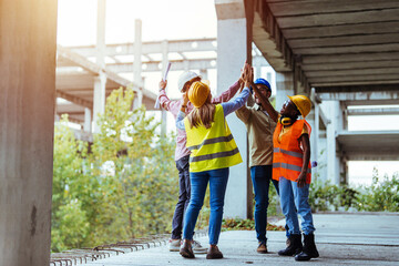 A group of diverse construction workers high-five each other while wearing safety helmets and...