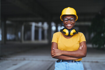 A smiling black woman in a yellow hard hat and safety glasses stands with crossed arms at a construction site, embodying professionalism in her field.