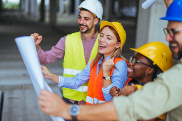 A group of cheerful construction professionals of diverse ethnicities review blueprints on-site,...