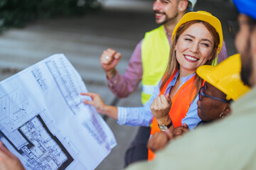 A group of joyful male and female construction workers in safety gear examine building plans...