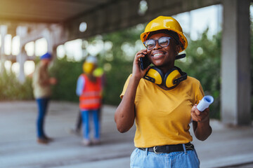 A cheerful black woman in a yellow hard hat engages in a phone conversation at a construction site,...