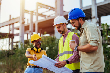 Three construction professionals in safety gear engage in discussion over a blueprint at a building site, reflecting collaboration in a diverse work environment.