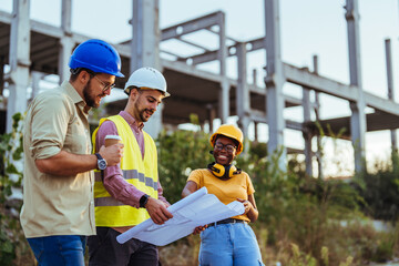A group of male and female construction professionals in hard hats and reflective vests actively discuss blueprints outdoors, with steel structures in the background.