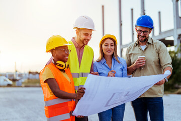 A group of focused construction professionals, featuring a blend of ethnicities and genders, review a large blueprint at a building site, clad in vibrant safety gear under fading daylight.