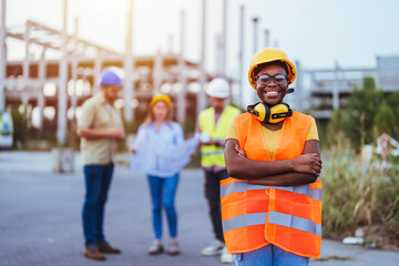 A smiling Black woman in safety gear stands with arms crossed at a construction site, her diverse...